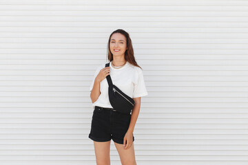 Woman, wearing white t-shirt, black shorts, fanny pack or waist pack and gold chain necklace, standing outdoor near white wall. Stylish trendy basic minimalistic casual outfit. Street fashion.