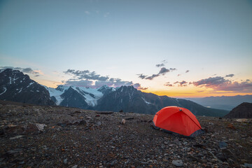 Scenic alpine landscape with tent at very high altitude with view to large mountains in orange dawn sky. Vivid orange tent with awesome view to high mountain range under cloudy sky in sunset colors.