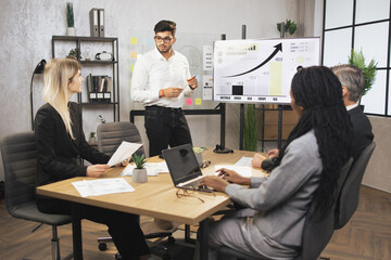 Serious indian businessman manager stands near big digital wall screen with company financial and growth charts and talks to his multiethnic colleagues, discussing results of the project.