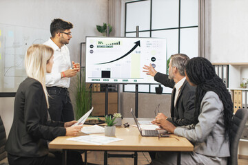 Business concept. Successful confident experienced diverse team of corporative workers, listening indian male expert, showing strategy of company, financial reports on the wall TV screen.