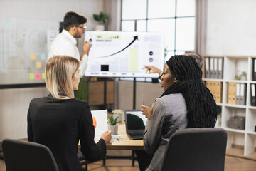 Two female african american and caucasian business womans, listening report of their indian male colleague, standing in front of the big plasma digital wall screen with infographics charts.