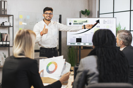 Middle Aged Male Bearded Indian Chief Showing Company's Work Infographics On Digital Wall Screen During Meeting With Team Of Diverse Multiracial Male And Female Businesspeople.