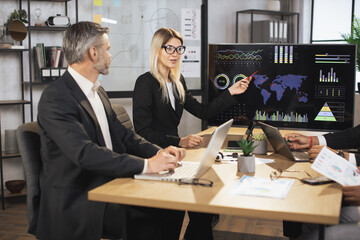 Board of diverse multiethnic directors business people at annual meeting in office room, listening mature blond lady and discussing work results. TV screen with map and charts on the background.