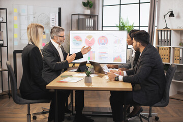 Team of diverse attentive mixed race business people listening their middle aged male bearded chief, showing dynamics of key ecological global problems, or world market, using digital wall screen.