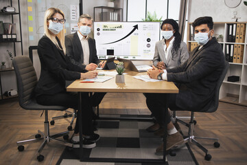 Multiethnic coworkers in modern office, wearing facial protective masks, working during covid-19 pandemic, sitting around the table and discussing the project.