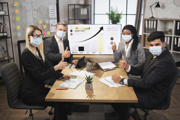 Work during covid pandemic. Happy multiethnic business people in face masks, showing thumbs up as agreement and teamwork, posing to camera in modern conference room in office.