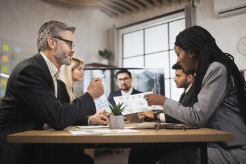 Fototapeta na wymiar Concentrated diverse businesspeople, having telemeeting at office in front of plasma TV screen. Young African woman discussing financial report with mature male bearded Caucasian colleague.