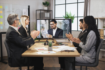 Group of four skillful multiethnical businesspeople clapping hands and looking at screen, having video conference in boardroom, with their male colleague explaining some financial charts.