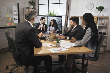 Focused multiracial business people in conference room, looking at a screen, having video conference with another team, African lady and Caucasian man, using smart video technology to communicate.