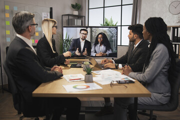 Focused multiracial business people in conference room, looking at a screen, having video conference with another team, African lady and Caucasian man, using smart video technology to communicate.