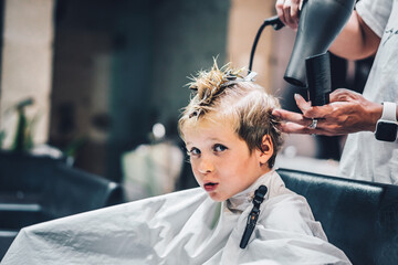 Naklejka premium Lifestyle photo Barber shop blond boy customer. Hairdresser man work move hand comb tool scissors dryer clip white cape. Modern style kid short hair cut. Fashion business service for children