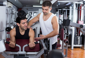 Male trainer explaining to muscular guy technique of performing strength exercises in gym