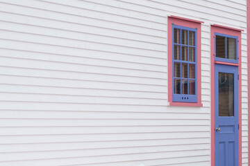 Fototapeta premium The exterior wall of a white wooden cape cod clapboard siding house with a purple panel door and vintage glass window, black metal hinges, pink trim around the door, and white snow on the step. 