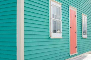 The exterior of a vibrant orange color single traditional wooden shutter door with cream color trim on a teal green clapboard wall of a historic house with four single hung windows with lace curtains.