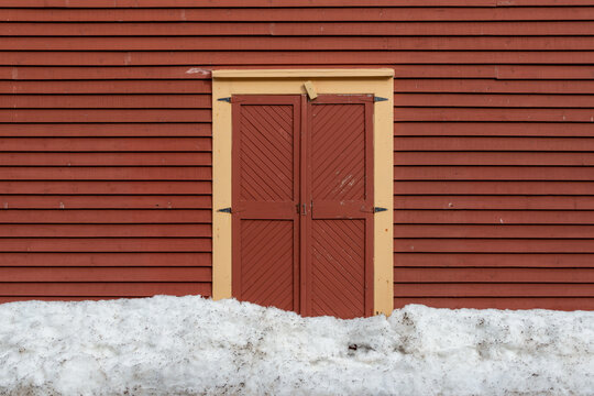 The exterior of a vintage rusty red color wooden wall of a rustic building. It has a yellow trim around the double wooden door. The wall is covered in narrow wood clapboard siding.  - Powered by Adobe