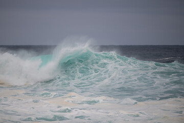 An angry turquoise green color massive rip curl of a wave as it barrels rolls along the ocean. The white mist and froth from the wave are foamy and fluffy. The ocean in the background is deep blue. 