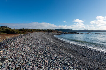 A sheltered pebbly beach on a bright sunny day. The sky is blue and there are white clouds in the background. A small wave rolls in on shore and there's ice and snow on some of the rocks and trees.