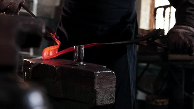 The blacksmith takes out heated oblong piece of metal and begins to bend in using forceps