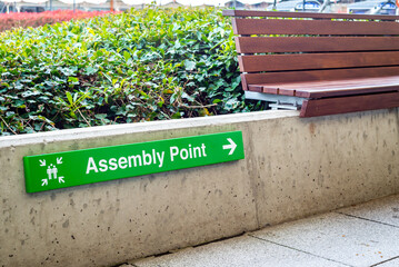 Green assembly point sign with people icon and arrow on wall beside wooden bench at shopping centre.