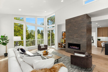 Beautiful and bright living room in new contemporary style luxury home ,with large fireplace and surround, wall of windows showcasing exterior view of trees and blue sky.
