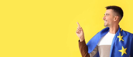Young man with flag of European Union pointing at something on yellow background with space for text