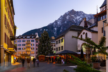 Evening view of central street in small mountainous Swiss town of Brig-Glis with main Christmas tree decorated with traditional lights with snow-capped Alpine peaks in background