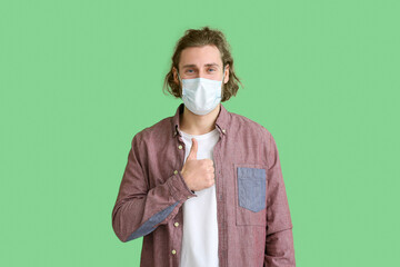 Young man wearing medical mask and showing thumb-up on green background