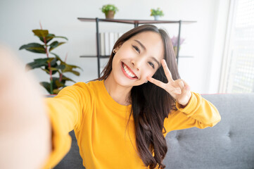 Pretty young asian female with big smile sitting at living room. She having fun taking light cheerful selfie on blurred background - Powered by Adobe