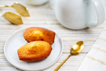 Madeleine cookie or French sponge cake in a white plate alongside white teapot, golden spoon and tong by a white napkin on white wood background.