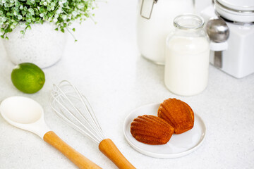 Madeleine cookie or French sponge cake in a white plate alongside white kitchen utensils, lime, sugar, milk and flour jars on white background.