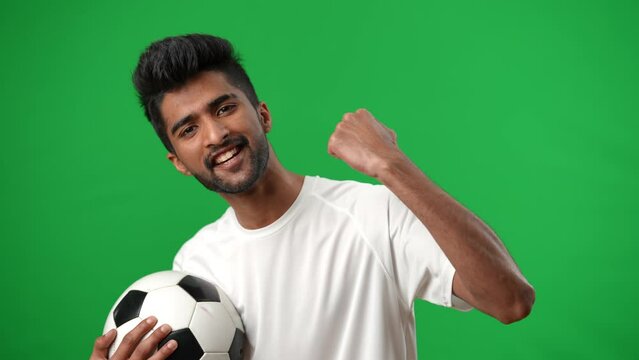 Inspired Middle Eastern man gesturing yes posing at chromakey background with football ball. Portrait of smiling young footballer looking at camera smiling on green screen