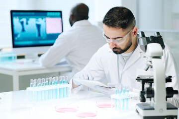 Contemporary young male scientist in workwear using digital tablet while searching for online data about new biotechnologies