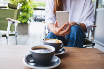 Closeup image of a young woman holding and using mobile phone while drinking coffee with friend in cafe