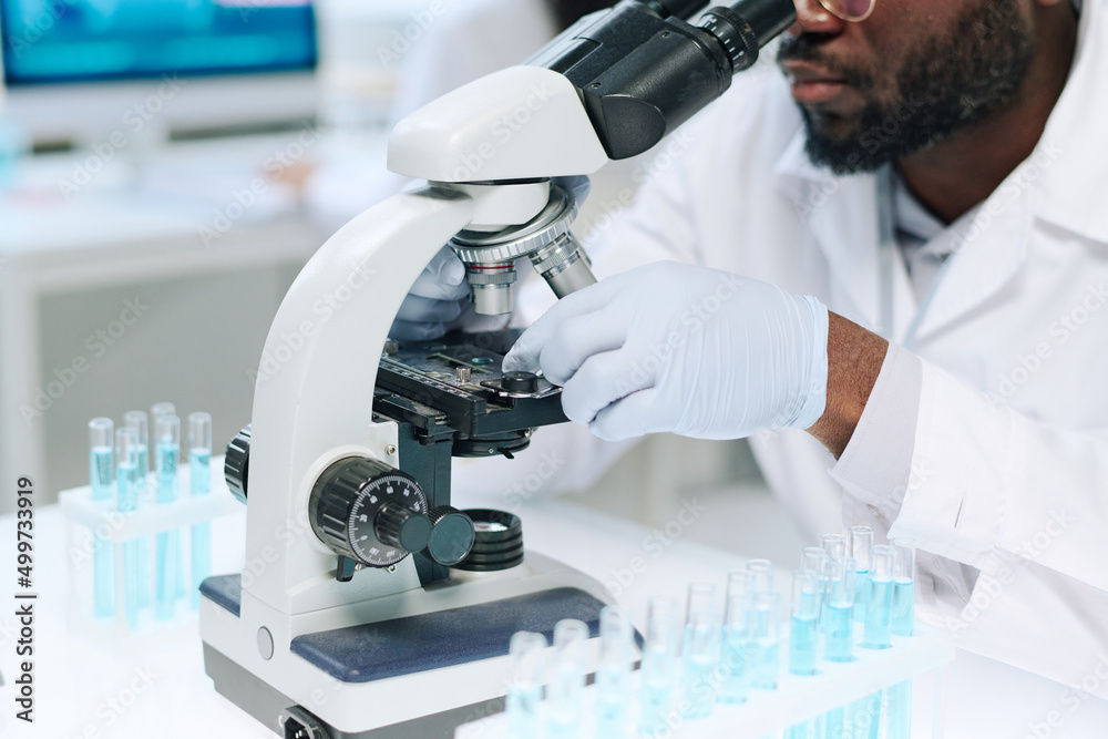 Poster gloved hands of modern african american male scientist studying new virus in microscope while sittin