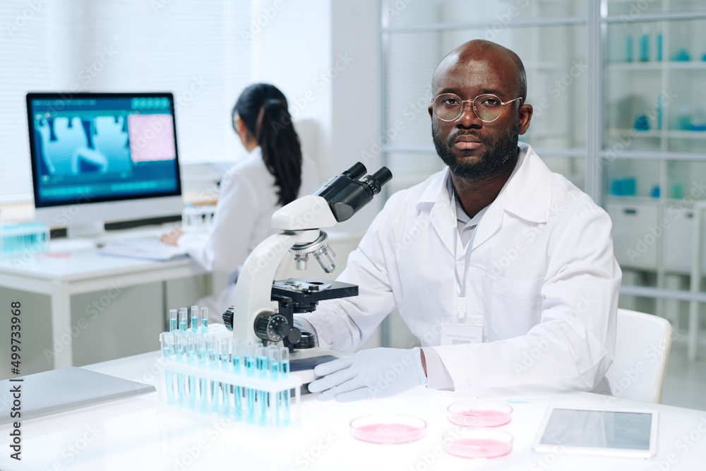 Poster Young serious scientist in lab coat sitting by workplace in front of microscope and looking at camera during scientific work