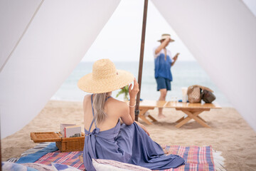 Woman wearing blue dress and straw hat and her boyfriend sitting under the white tent on a beach.