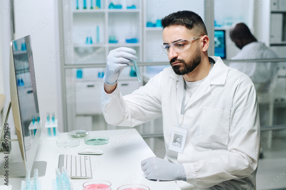 Poster young serious male microbiologist or virologist studying blue liquid in flask while sitting by desk 