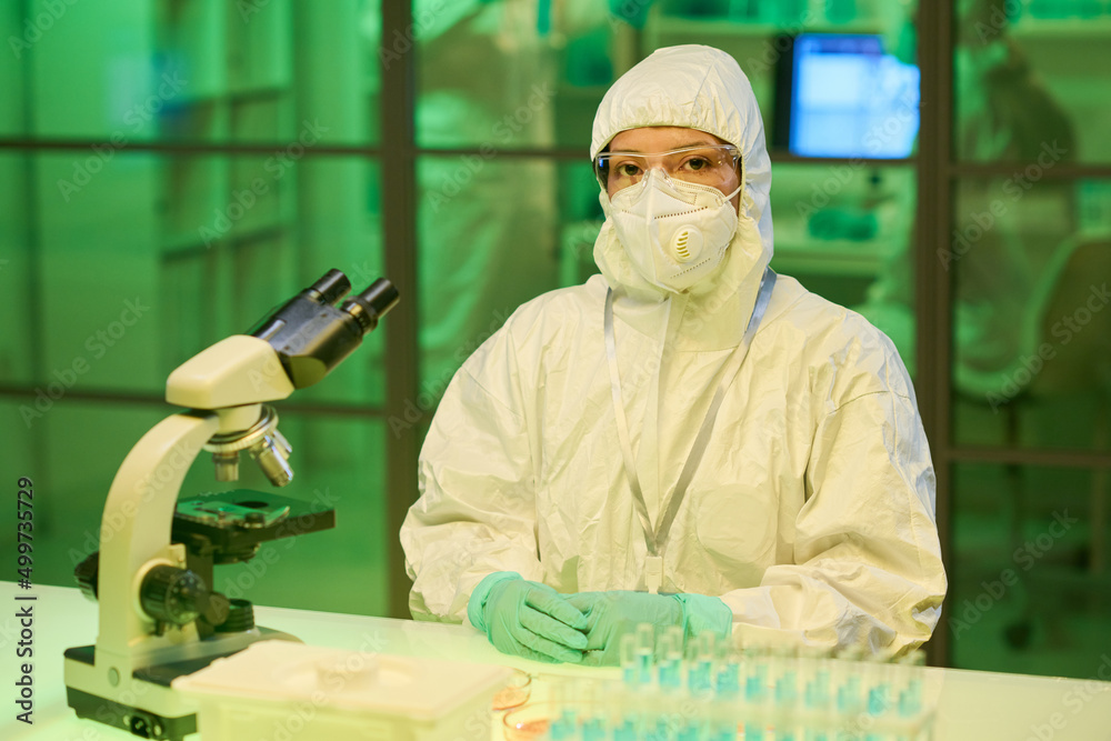 Poster young contemporary scientist in biohazard suit sitting by workplace with microscope and group of fla