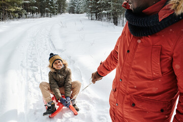 Happy cute little boy in winterwear looking at his father pulling sledge with him while moving...