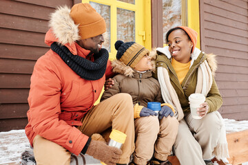 Cheerful black woman in winterwear looking at her cute son during talk while sitting on porch of cottage next to young man and having tea