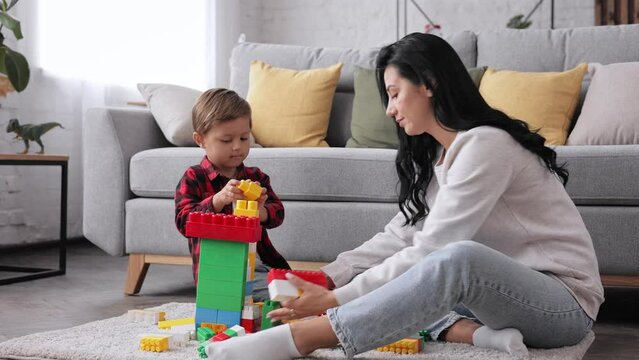 Family spending free time at home. Mom sitting on floor with little kid teaches son to build tower of colored blocks. Mother teach kid, favourite hobby and leisure concept. Mom playing with small kid.