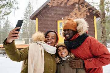 Happy family posing for selfie against country house on snowy winter day while young black woman with smartphone taking shot