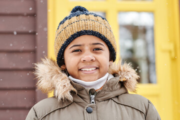 Happu adorable boy in warm winter jacket and striped knitted beanie hat looking at camera while standing against yellow door of country house