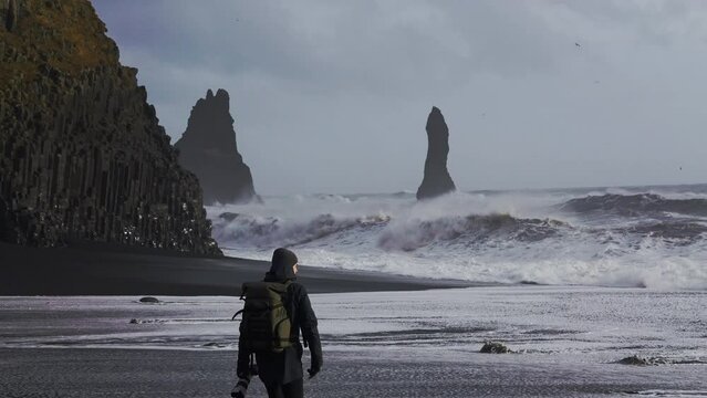 Photographer Walking Along Black Sand Beach To Photograph Rough Sea
