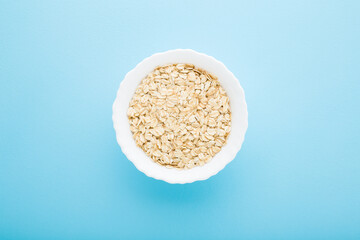 White bowl with dry rolled oat on light blue table background. Pastel color. Closeup. Healthy food. Top down view.