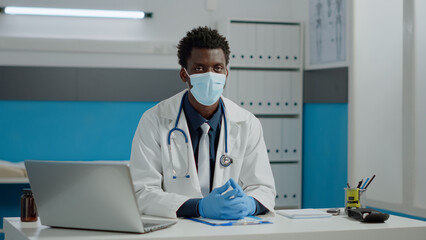 Portrait of african american doctor wearing face mask and white coat with stethoscope in medical cabinet. Black young medic looking at camera while sitting at desk in healthcare office