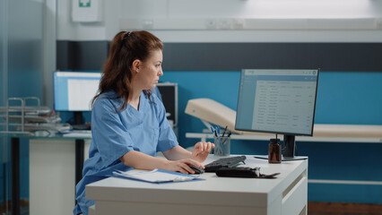 Medical assistant working on computer with patient information at desk. Woman nurse using keyboard and monitor in cabinet while checking healthcare documents and papers for appointments.