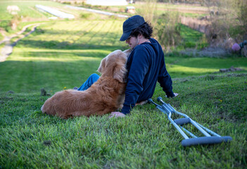 Big Golden Retriever dog cuddles in grass with young man and crutches