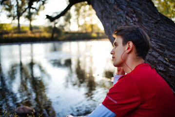 Young sportsman listening music, while sitting and resting outdoors near river.