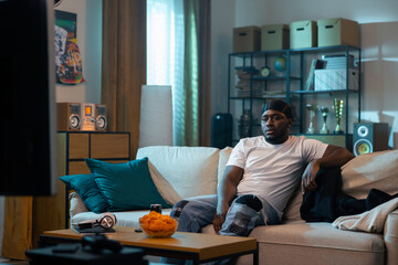 A bored guy is relaxing on the living room sofa in front of the television. The young man stares at a weak movie, TV series, comedies, losing soccer game. On the table bowl of chips and canned drink.